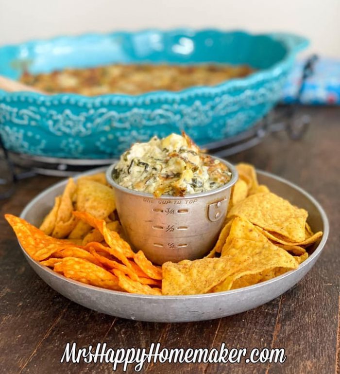 Spinach artichoke alfredo dip in a blue oval casserole dish With a round dip tray with chips beside it 