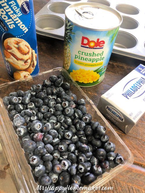 Fresh blueberries, a package of cinnamon rolls, a can of crushed pineapple, and a package of cream cheese on the counter with a muffin cup behind them