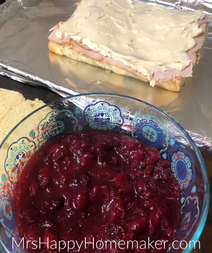 cranberry sauce in a bowl beside of the baked sliders being prepped - bread, meat, and cheese 