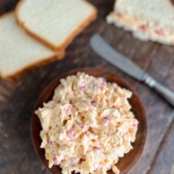 Pimento cheese in a brown bowl on a table with a knife & two slices of bread