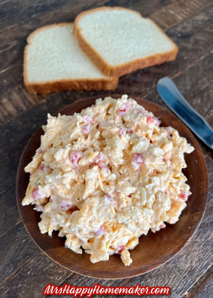 Pimento cheese in a brown bowl on a table with a knife & two slices of bread