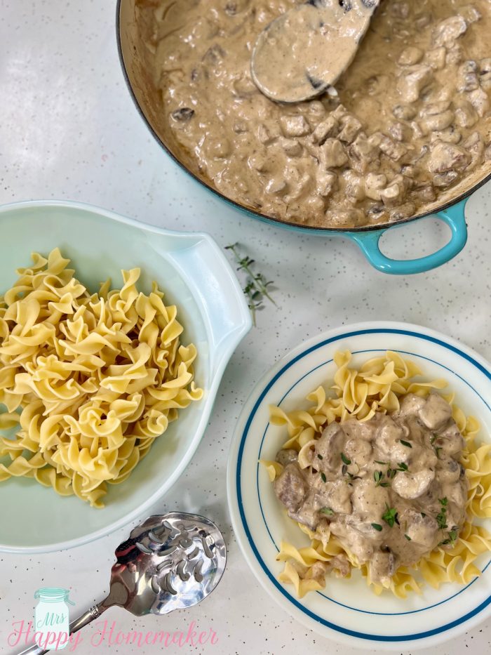 Beef stroganoff over noodles in a white bowl with a blue rim. Beside it is a white bowl full of plain noodles and a blue skillet full of the beef mixture 