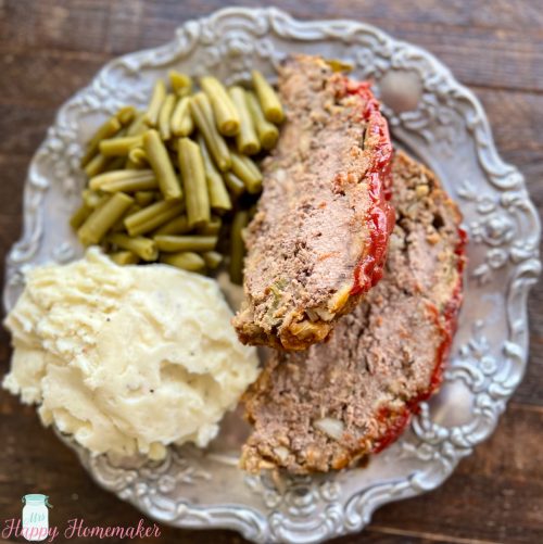 Two slices of meatloaf on a pewter plate, surrounded by mashed potatoes & green beans
