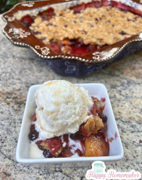 Peach Blueberry Crisp in a 9x13 brown and blue casserole dish With a serving in a small white dish & ice cream on top