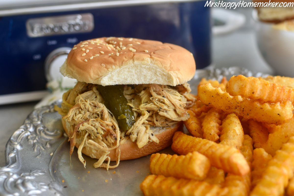 Dill Pickle chicken in a crockpot and piled on a bun thats on a plate beside the crockpot