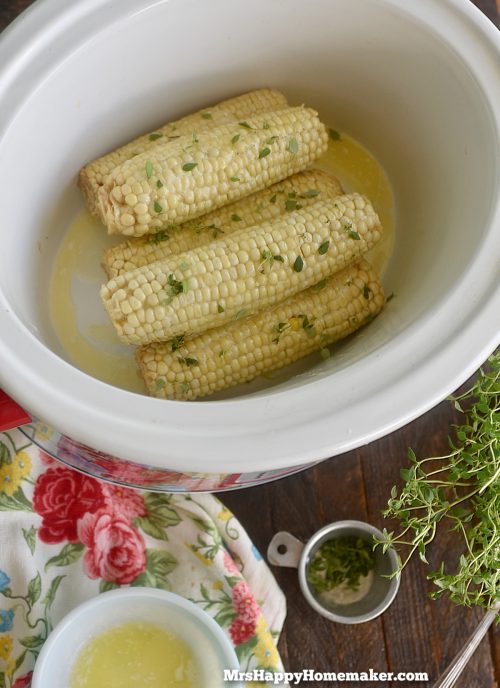 Corn on the cob inside of a white crockpot surrounded with a white bowl of melted butter and fresh herbs
