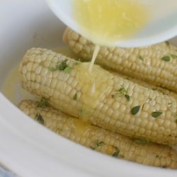 Corn on the cob in a white crockpot with melted butter being poured on top from a white bowl