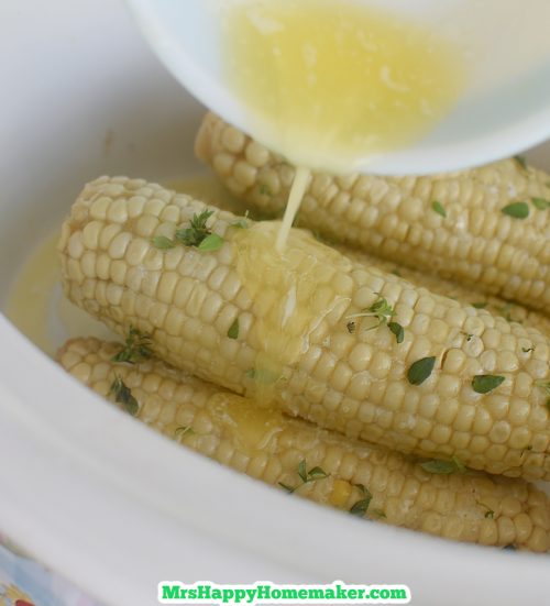 Corn on the cob in a white crockpot with melted butter being poured on top from a white bowl