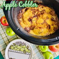 Apple cobbler in a black crockpot with an ‘apple pie’ design plate beside of it
