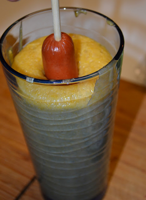 A corn dog being dipped into a large blue glass full of batter