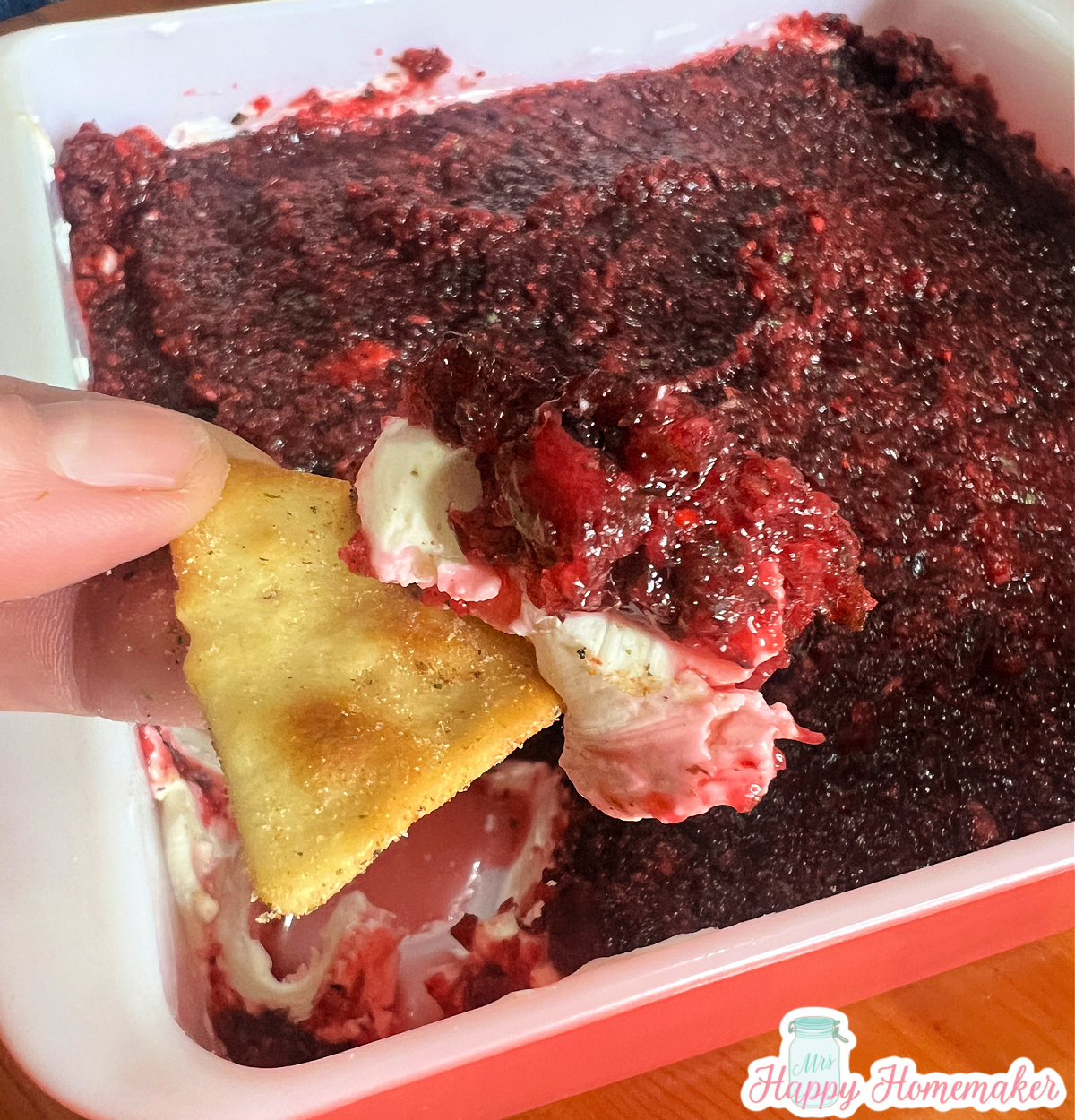 Cranberry jalapeno roomkaasdip op een cracker met daaronder een roze vierkante schaal vol dip 