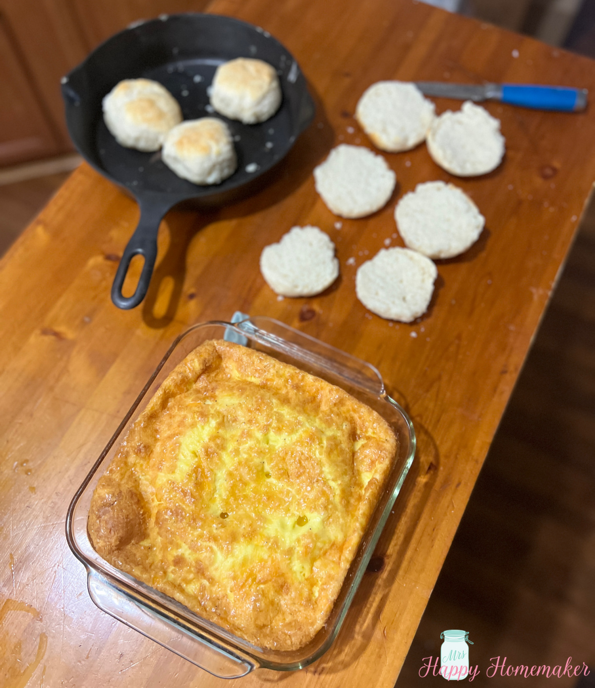 Baked Eggs inside of a square clear baking dish beside of a cast iron skillet with biscuits on a kitchen island with a few split biscuits on the counter