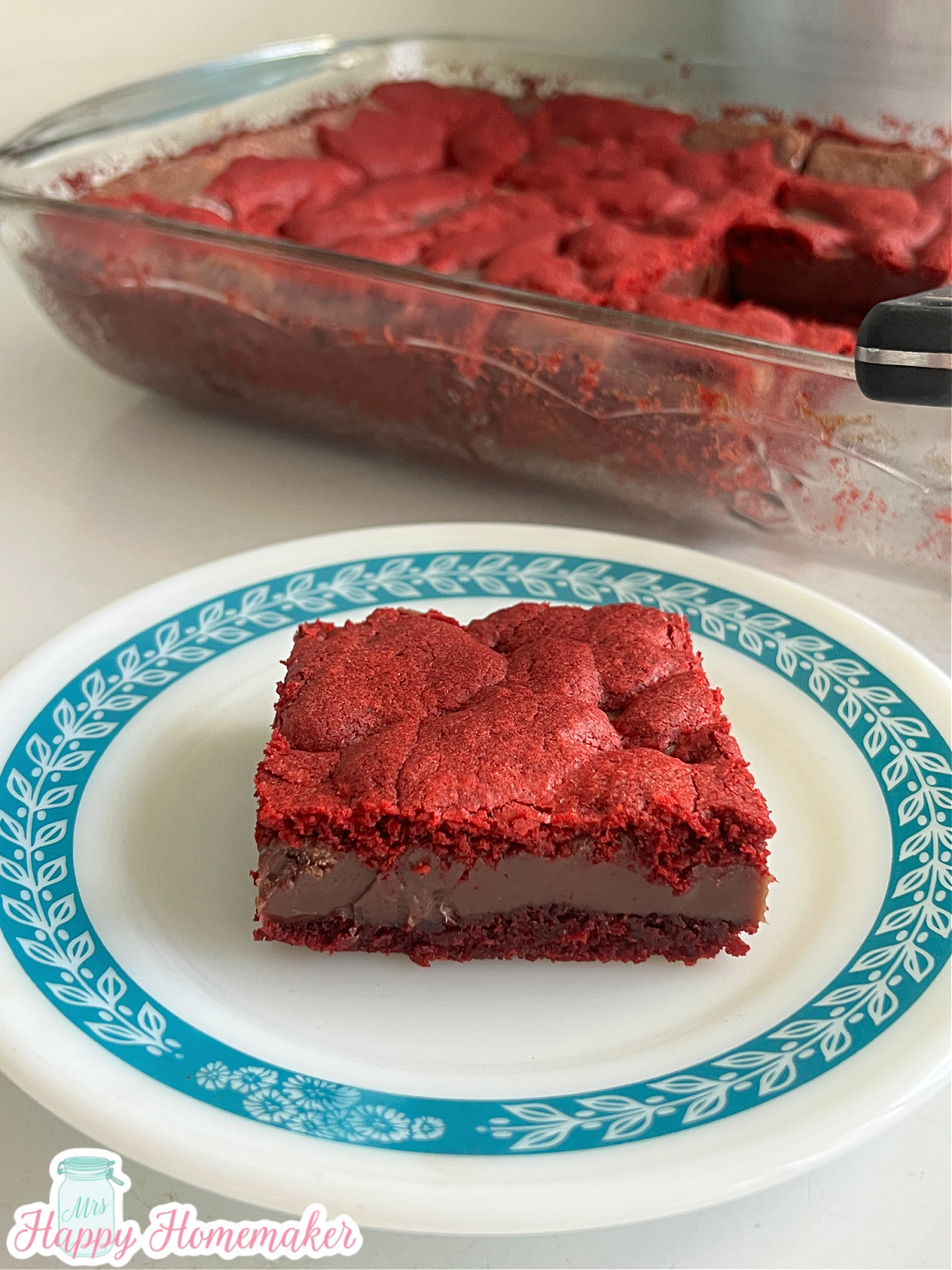 red velvet fudge bar on a blue rimmed white plate on a white counter with a clear casserole dish behind it with the rest of the bars