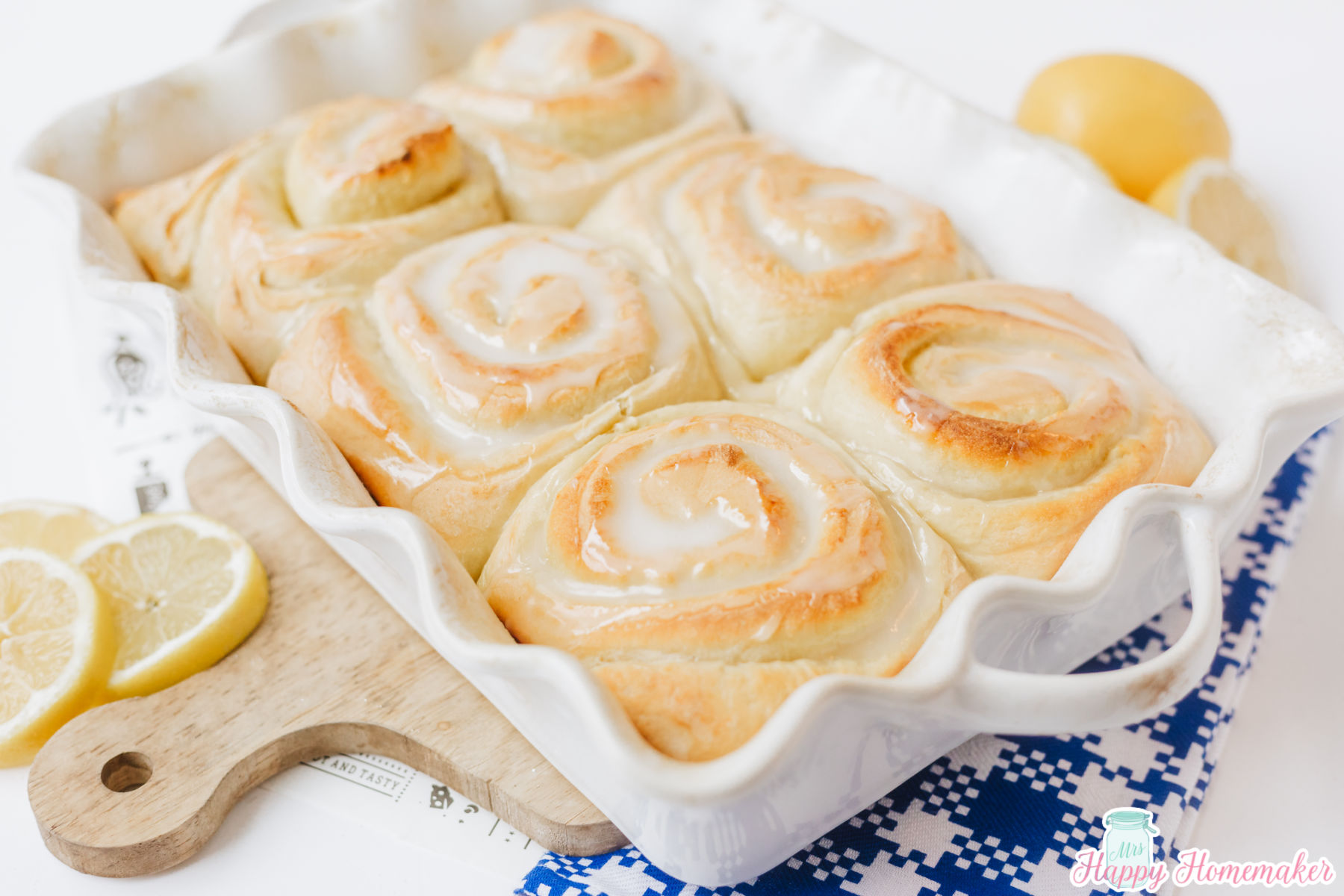 Iced Lemon Rolls in a 9x13 ruffled white baking dish with some lemon garnishes on the counter around it